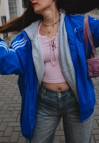 Μπλε αθλητικό μπουφάν πάνω από ένα ανοιχτό γκρι φούτερ, συνδυασμένο με ροζ ριμπ ωμοποιήτη και τζιν ψηλής μέσης σε ανοιχτό χρώμα. Μικρή μοβ τσάντα χεριού.