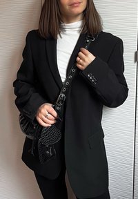 Femme portant un blazer noir et un col roulé blanc tenant un sac à main noir texturé avec une sangle  à boucle contre un mur blanc texturé.