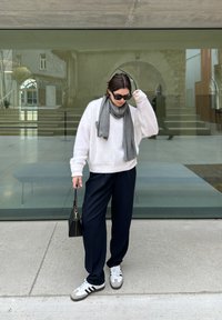 Hvid, fluffy sweater, grå tørklæde, marineblå vide bukser, sort håndtaske og grå/hvide sneakers, stående foran glasarkitektur.