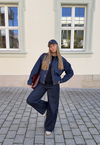 Chaqueta de denim y jeans acampanados en azul oscuro, combinados con una blusa negra. Los accesorios incluyen una gorra oscura y un bolso burdeos. Fondo de piedra pavimentada.