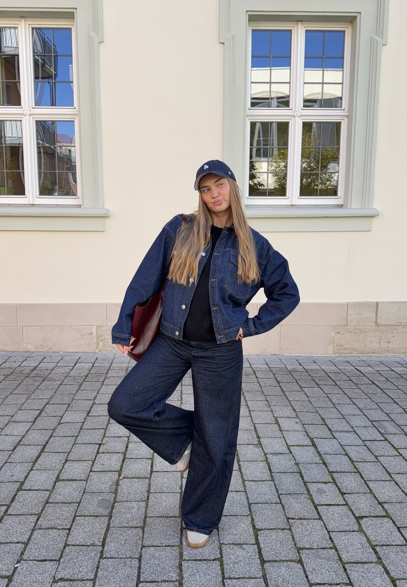 Denimjakke og flagrende jeans i mørkeblå, parret med en sort top. Accessories inkluderer en mørk hat og en bordeaux taske. Belægning af sten i baggrunden.