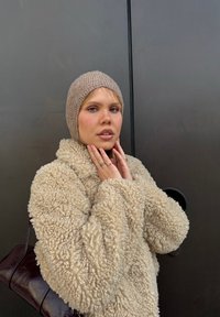 Beige fuskpälsjacka med en texturerad yta, bärs över en åtsittande beige stickad mössa. Bakgrunden har en slät mörk yta och ett cirkulärt detalj.