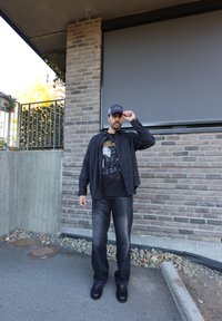 Black check shirt worn over a black graphic t-shirt, teamed with dark jeans and black shoes. Standing against a stone wall backdrop.