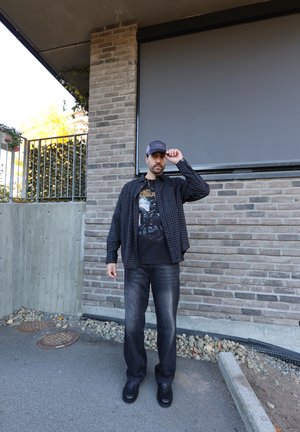 Camisa de cuadros negra sobre una camiseta gráfica negra, combinada con jeans oscuros y zapatos negros. De pie frente a un fondo de pared de piedra.