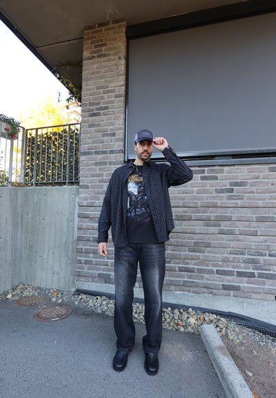 Camisa de cuadros negra sobre una camiseta gráfica negra, combinada con jeans oscuros y zapatos negros. De pie frente a un fondo de pared de piedra.