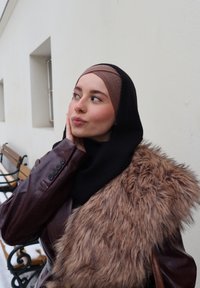 Jeune femme en veste en cuir marron avec bordure en fausse fourrure et foulard, de profil, reposant sa main sur sa joue à l'extérieur près de bancs.