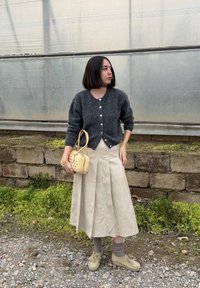 Femme aux cheveux courts et foncés portant un cardigan gris, une jupe plissée beige, des chaussures beiges, et tenant un petit sac à main beige, debout en plein air sur du gravier.