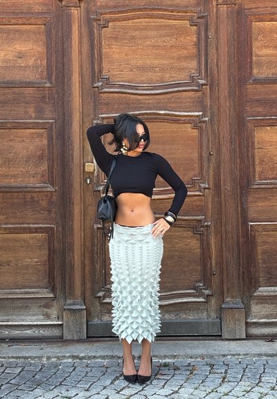 Falda gris asimétrica con textura de picos en 3D, combinada con una blusa negra ajustada de manga larga. Tacones altos negros y un bolso pequeño completan el look.