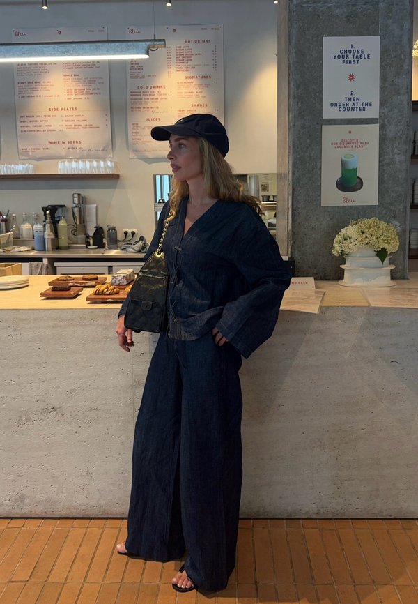 Femme en tenue bleu foncé et casquette noire se tient au comptoir du café avec des pâtisseries, des tableaux de menu et un vase à fleurs derrière elle.