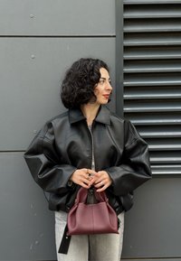 Giacca in pelle nera con zip e maniche ampie, abbinata a una piccola borsa bordeaux e jeans grigi chiari, su uno sfondo grigio texturizzato.