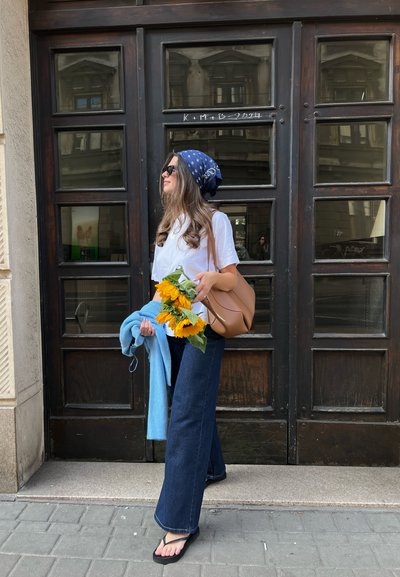 Una mujer se encuentra cerca de una puerta de madera, vistiendo una camisa blanca, un pañuelo azul en la cabeza, jeans oscuros de campana, chanclas y sosteniendo girasoles con una bolsa marrón.