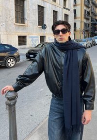 Veste en cuir noir, écharpe bleu foncé et jean bleu. La veste a une texture lisse, tandis que l'écharpe est douce avec des extrémités à franges. Cadre urbain.
