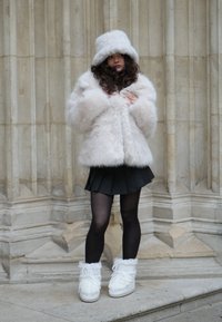 Kremni plašč iz umetnega krzna s klobukom v obliki vedra v enaki barvi, črna nabrana krila, prozorne črne nogavice in bele škornje z umetnim krznom ob strani ter zavezovanjem.