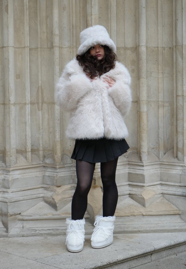 Tekoturkisinen kermaisen värinen takki ja siihen sopiva lierihattu, musta laskostettu hame, läpikuultavat mustat sukkahousut ja valkoiset nauhalliset turkiskaistaleisilla saappailla.