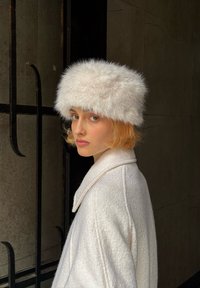 Λευκό καπέλο από γούνα με φουντωτή υφή και στρογγυλό σχήμα. Το μοντέλο φοράει ένα κρέμα χρώματος παλτό με φαρδύ γιακά. Στο φόντο υπάρχουν σκούρες γραμμές.