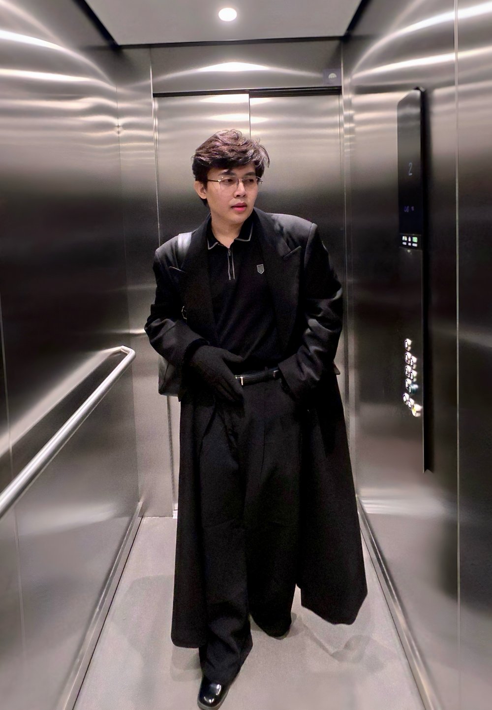 Jeune personne portant des lunettes, un long manteau noir, des gants noirs et un pantalon noir, se tenant à l'intérieur d'un ascenseur en acier inoxydable avec une rampe.