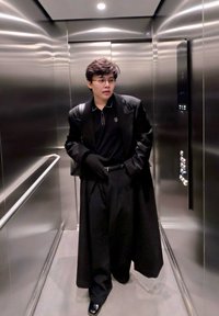 Jeune personne portant des lunettes, un long manteau noir, des gants noirs et un pantalon noir, se tenant à l'intérieur d'un ascenseur en acier inoxydable avec une rampe.