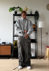 Sweat à capuche gris à fermeture éclair avec une coupe décontractée, associé à un pantalon cargo ample et sombre avec poches, et des baskets blanches sur un sol en bois clair.