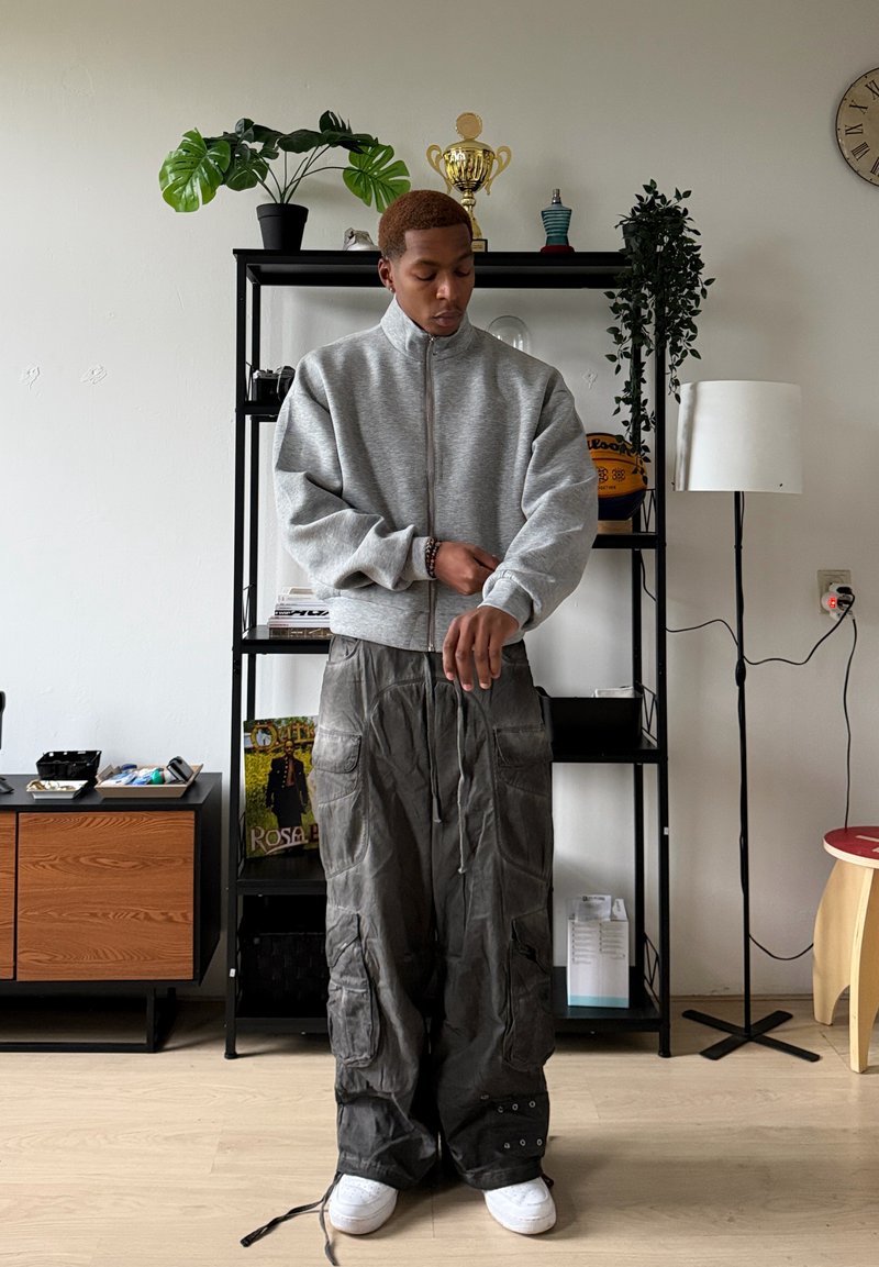 Sweat à capuche gris zippé avec coupe décontractée, associé à un pantalon cargo foncé et ample avec poches, et des baskets blanches sur un sol en bois clair.