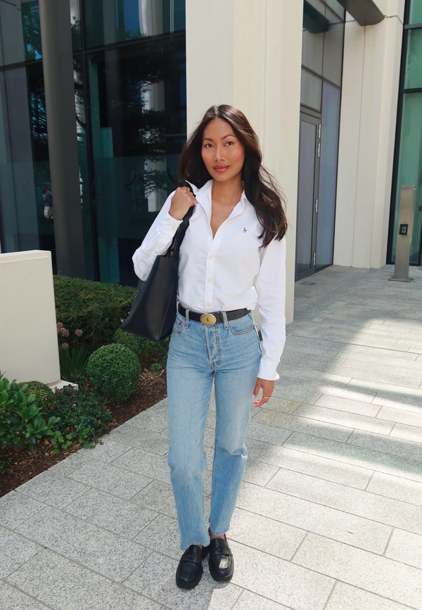 Jeune femme aux longs cheveux foncés portant une chemise blanche, un jean bleu, une ceinture noire et des chaussures noires, portant un sac fourre-tout noir à l'extérieur près d'un bâtiment moderne.
