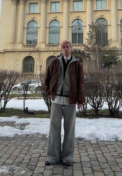Joven con cabello rosa, vestido con chaquetas superpuestas y pantalones anchos, está de pie en un camino empedrado cubierto de nieve frente a un gran edificio histórico.