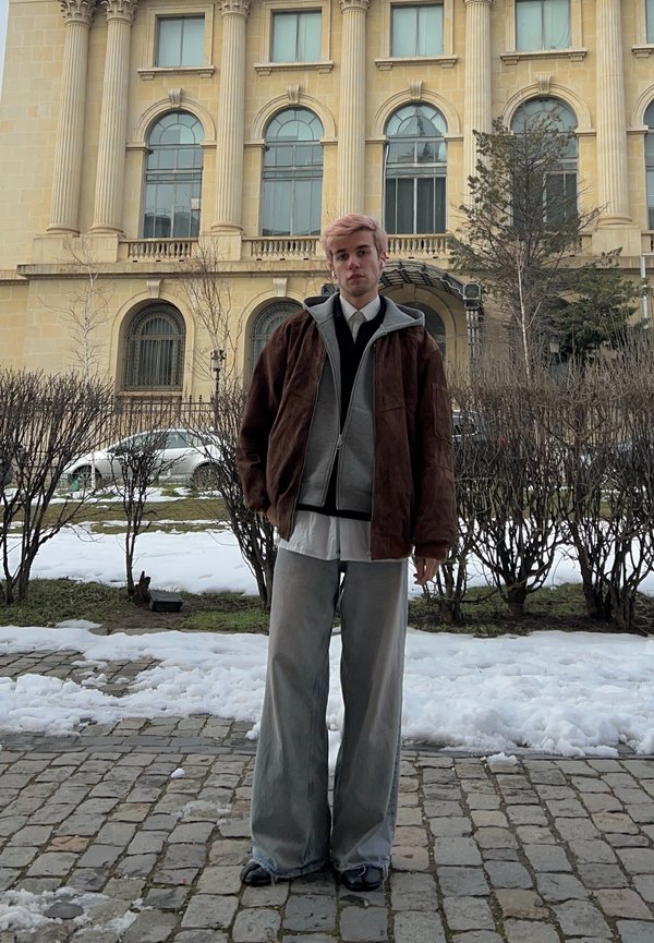 Rózsaszín hajú fiatal áll hóborította macskaköves úton egy nagy történelmi épület előtt, vastag kabátokat és bő szárú nadrágot viselve.