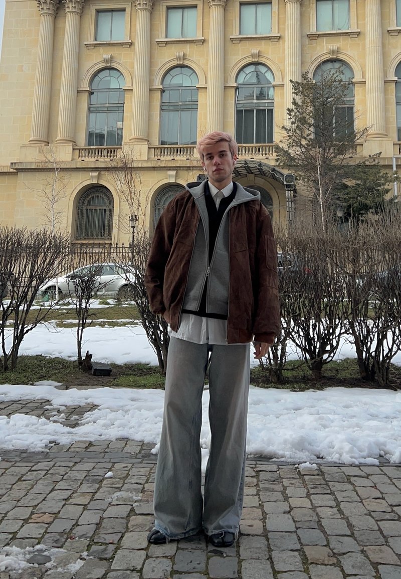Young person with pink hair, dressed in layered jackets and wide trousers, stands on a snowy cobblestone path in front of a large historic building.