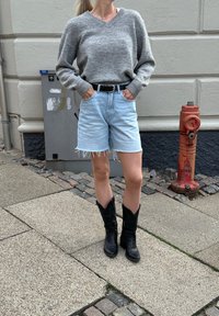 Siv svetrov iz pletenine, svetlo modri jeans kratki hlače s strganimi robovi, črn pas in črne kavbojske čevlje. Urbano okolje z pločnikom in hidrantom.