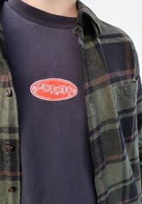 Chemise en flanelle à carreaux verts et noirs portée sur un T-shirt bleu marine orné d'un écusson ovale rouge avec des lettres blanches. Fermeture à boutons, texture douce.