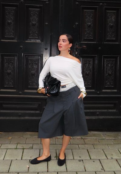 Top blanco de hombros descubiertos, pantalones culottes gris de pierna ancha, zapatos planos negros y bolso negro. Pulsera dorada como detalle. Puerta negra con textura de fondo.