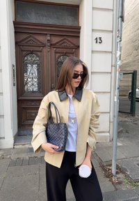 Jachetă bej cu fermoar și guler negru, tricou alb, pantaloni negri și o geantă țesută bleumarin. Cadru în aer liber cu o ușă din lemn în fundal.