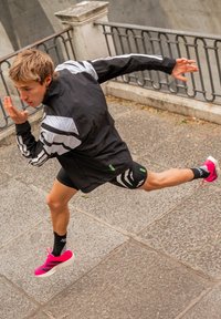 Ténis de corrida em rosa vivo com solas brancas, casaco leve preto com riscas cinzentas e calções pretos. Ambiente urbano com pavimento em pedra.