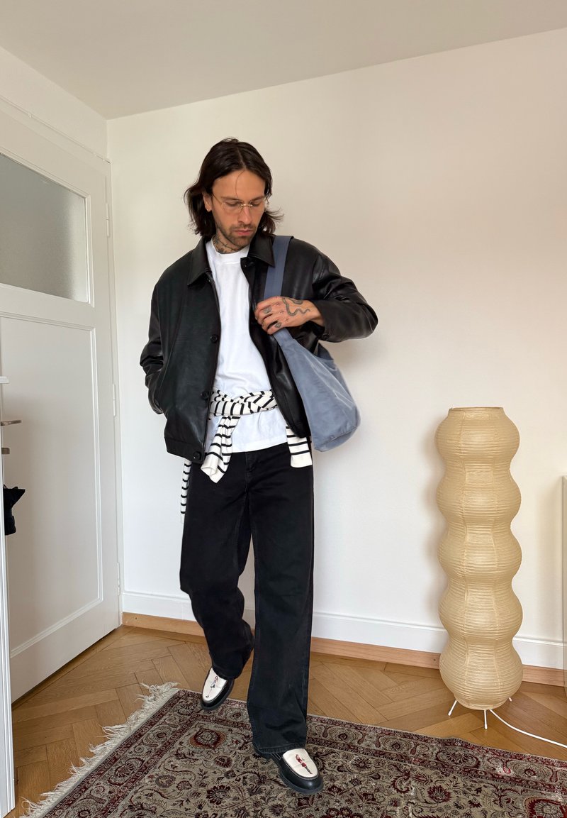 Man met zwarte leren jas, wit hemd, zwarte broek, gestreepte trui geknoopt rond de taille, draagt een blauwe tas, staat binnen op een patroonvloerkleed.