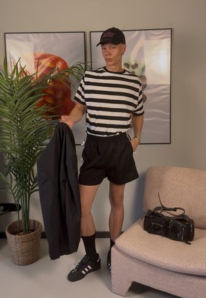 Camiseta de rayas blanca y negra, pantalones cortos negros, sosteniendo una chaqueta negra. Accesorios: gorra negra, bolsa negra y zapatillas negras con rayas blancas.