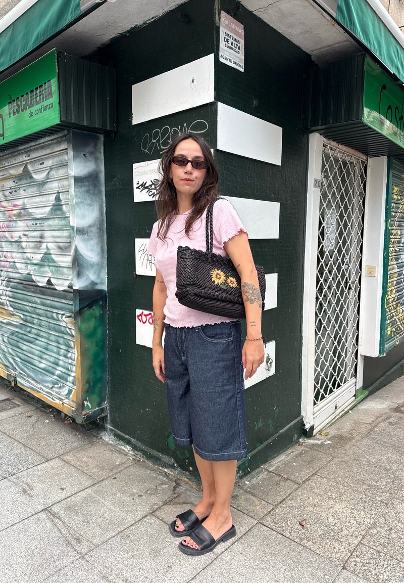 Junge Frau mit Sonnenbrille, pinkem Top, Jeans-Shorts, schwarzen Slides und schwarzer geflochtener Tasche mit Sonnenblumen steht an einer städtischen Gehweckecke.