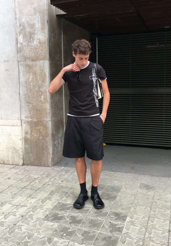 Jonge man staat buiten in zwarte shorts, zwart T-shirt met wit patroon, zwarte schoenen en houdt een zonnebril dicht bij zijn gezicht.