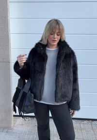 Femme aux cheveux blonds portant une veste en fourrure noire, un pull gris et un pantalon noir, tenant un sac à main noir à franges contre un mur blanc.