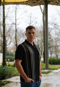 Svart kortärmad polo med vertikala grå ränder, lättviktigt tyg och ¼ dragkedja vid kragen. Kombinerad med blå jeans, stående utomhus.