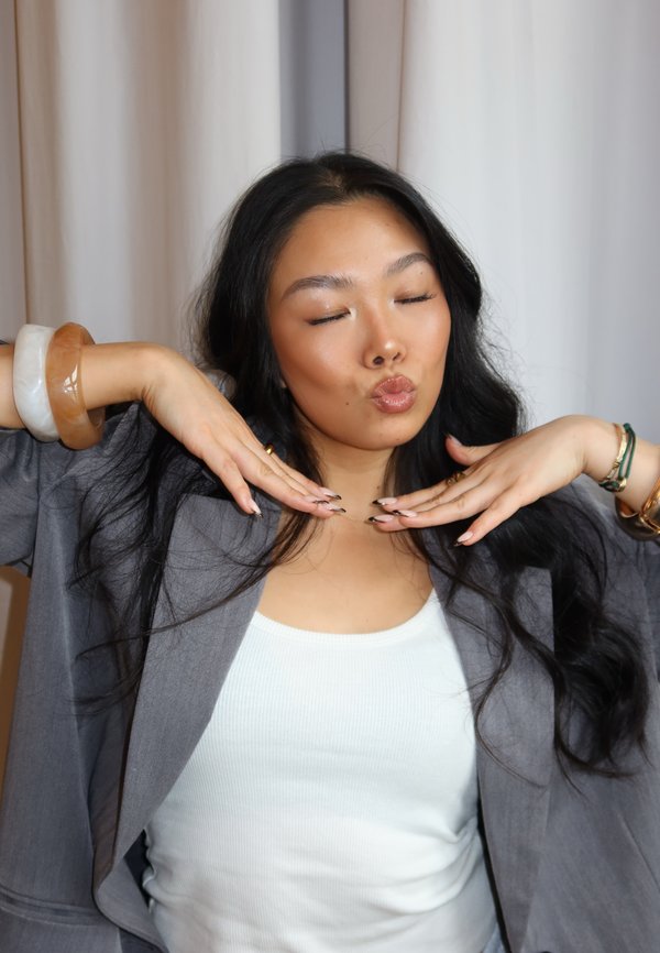 Blaser gris sur un débardeur blanc à côtes, accessoirisé avec de grands bracelets translucides. Cheveux longs et noirs et ongles manucurés dans une pose.