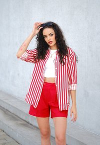 Camicia a righe rosse e bianche con maniche arrotolate, indossata sopra un crop top bianco e pantaloni shorts rossi a vita alta. L'outfit presenta un taglio moderno.