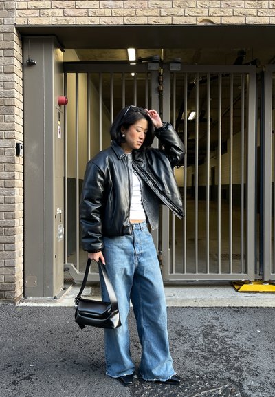 Mujer con chaqueta de cuero negra, camiseta blanca, vaqueros azules holgados, sosteniendo un bolso negro, de pie junto a una puerta metálica fuera de un edificio de ladrillo.