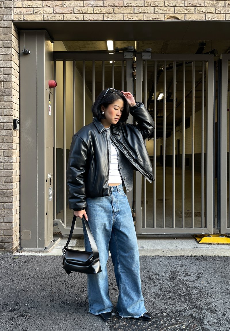 Femme en veste en cuir noire, haut blanc, jean bleu ample, tenant un sac à main noir, debout près d'une grille en métal devant un bâtiment en briques.