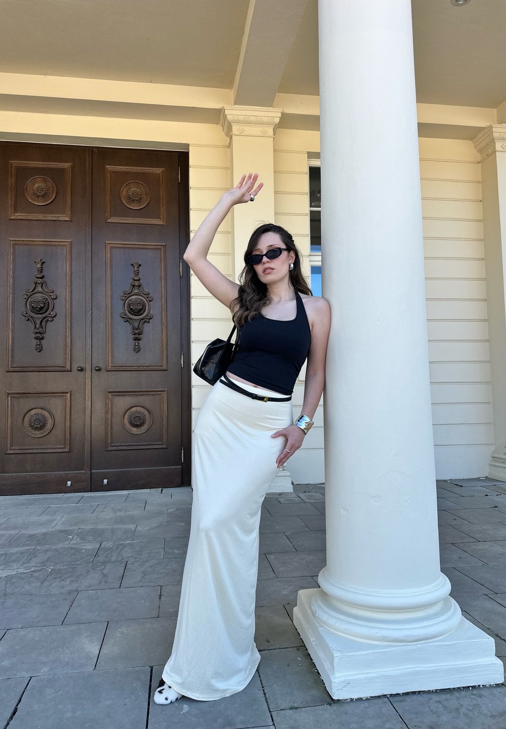 Black halter neck top, cream maxi skirt, sunglasses, and a black handbag. Standing next to a white column with wooden double doors in the background.