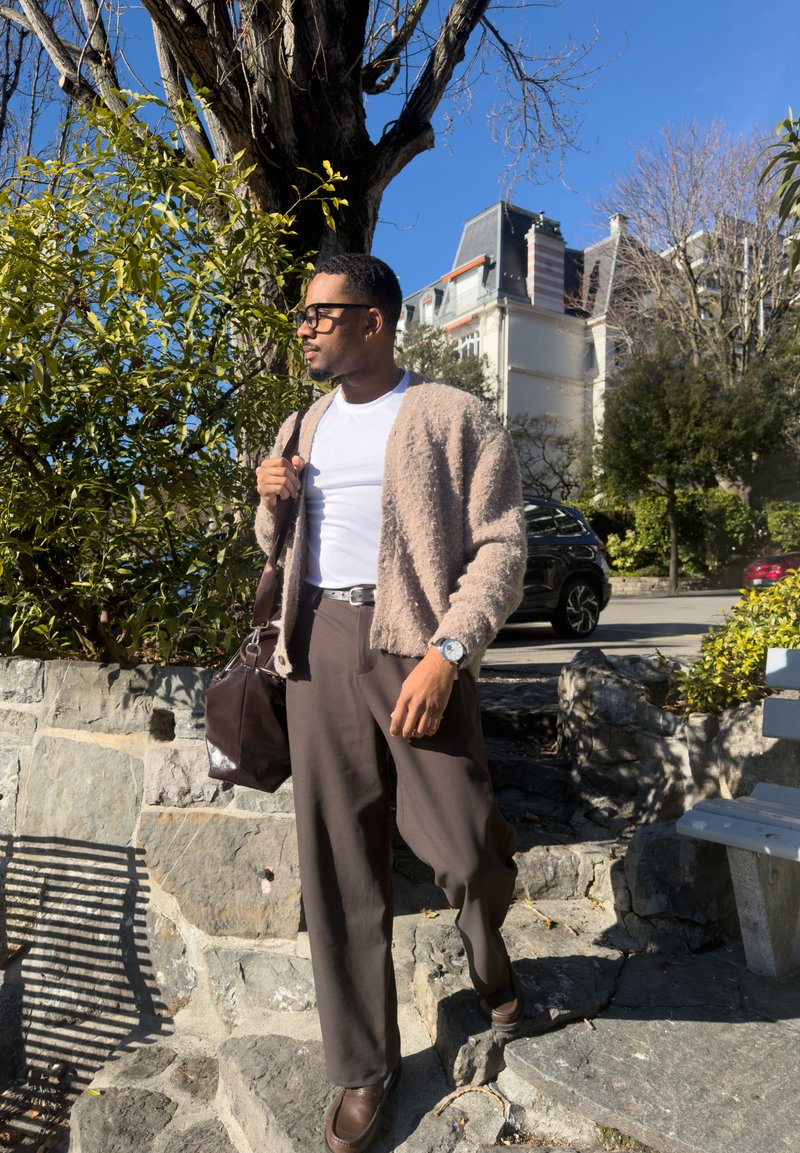 Homme portant des lunettes, chemise blanche, cardigan beige et pantalon marron, debout sur des marches en pierre près d'un arbre et de voitures garées par une journée ensoleillée.