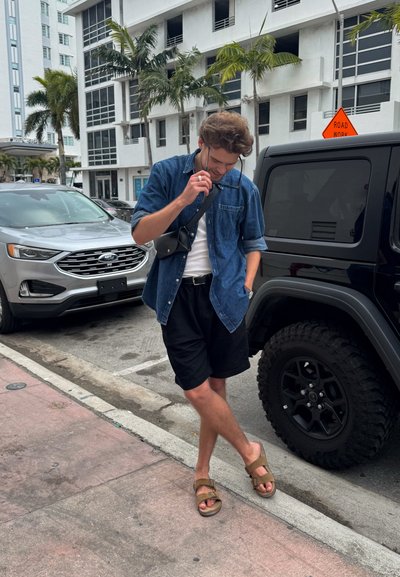 Una figura masculina se encuentra de manera casual en una acera, vistiendo una camisa de mezclilla, una camiseta blanca sin mangas, pantalones cortos negros y sandalias marrones, con gafas de sol en la mano.