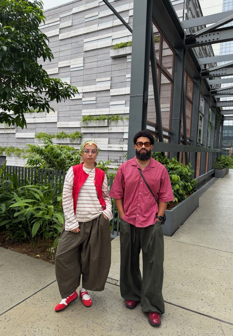 Deux personnes vêtues de pantalons amples et de hauts rouges se tiennent à l'extérieur près d'une végétation urbaine et d'un bâtiment gris moderne avec une structure métallique.