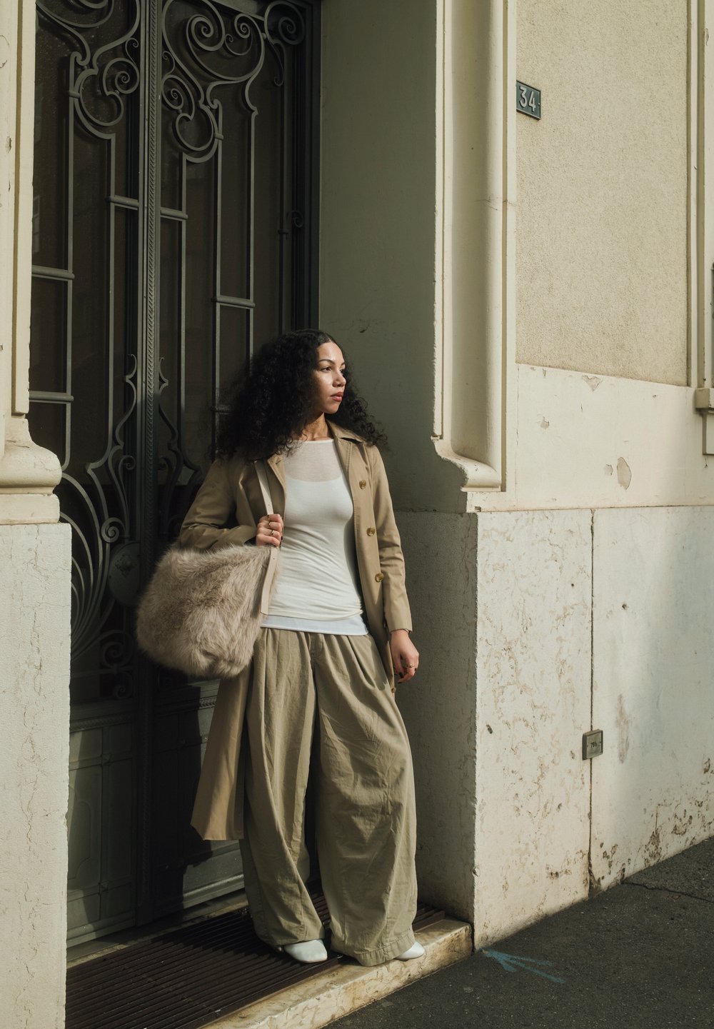 Beige trenčkot přes bílý top, široké béžové kalhoty a bílé boty. Drží chlupatou světle béžovou tašku. Pozadí obsahuje kovanou železnou bránu.