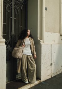 Beige trenčkot přes bílý top, široké béžové kalhoty a bílé boty. Drží chlupatou světle béžovou tašku. Pozadí obsahuje kovanou železnou bránu.