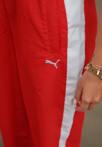 Røde treningsbukser har hvite sidestriper og en liten grå Puma-logo på venstre ben. Tøyet ser lett og glatt ut.
