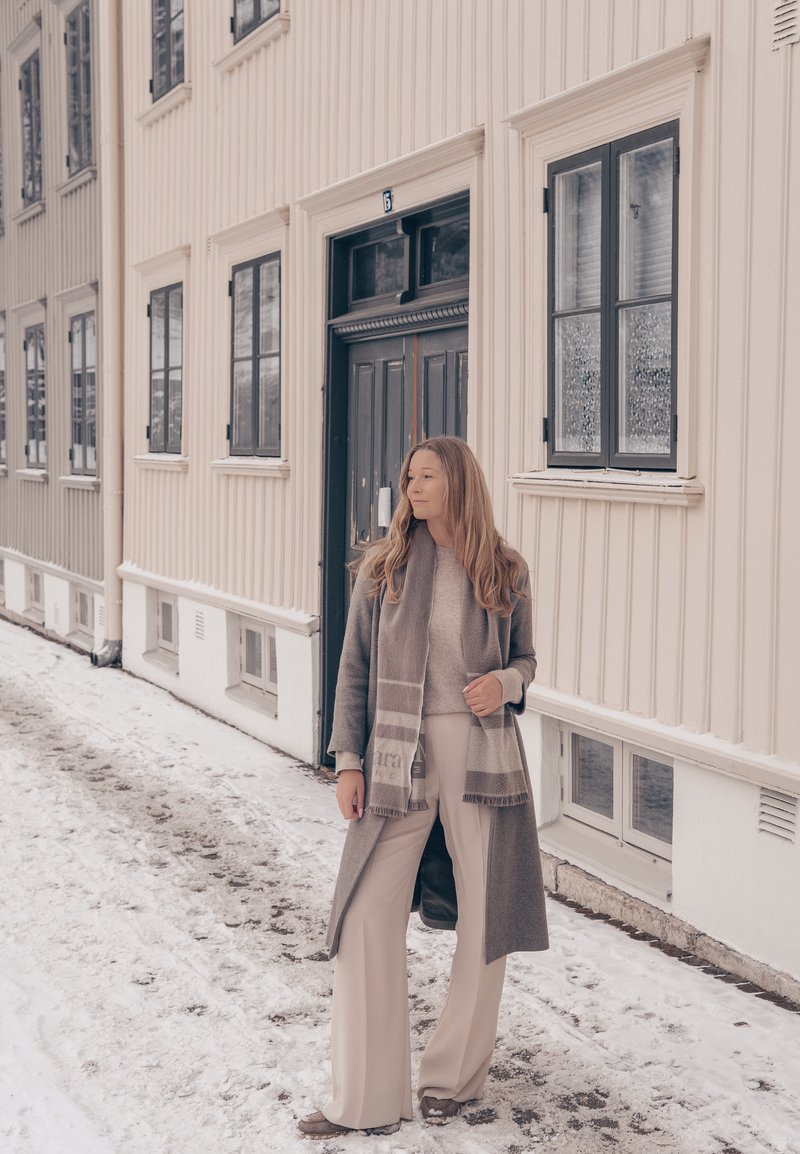 Kvinde iført en grå frakke og tørklæde, stående på en snedækket gade ved siden af en beige bygning med sortindrammede vinduer og dør.